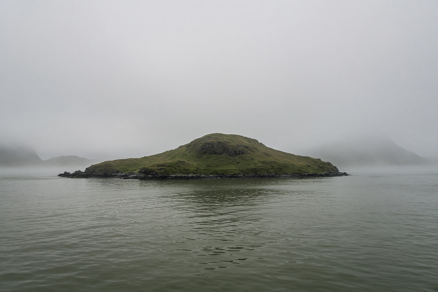 A small green island in grey-green water, seen from a boat.